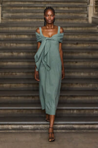 Model in a sage-green off-the-shoulder dress with a front knot standing on stone steps.
