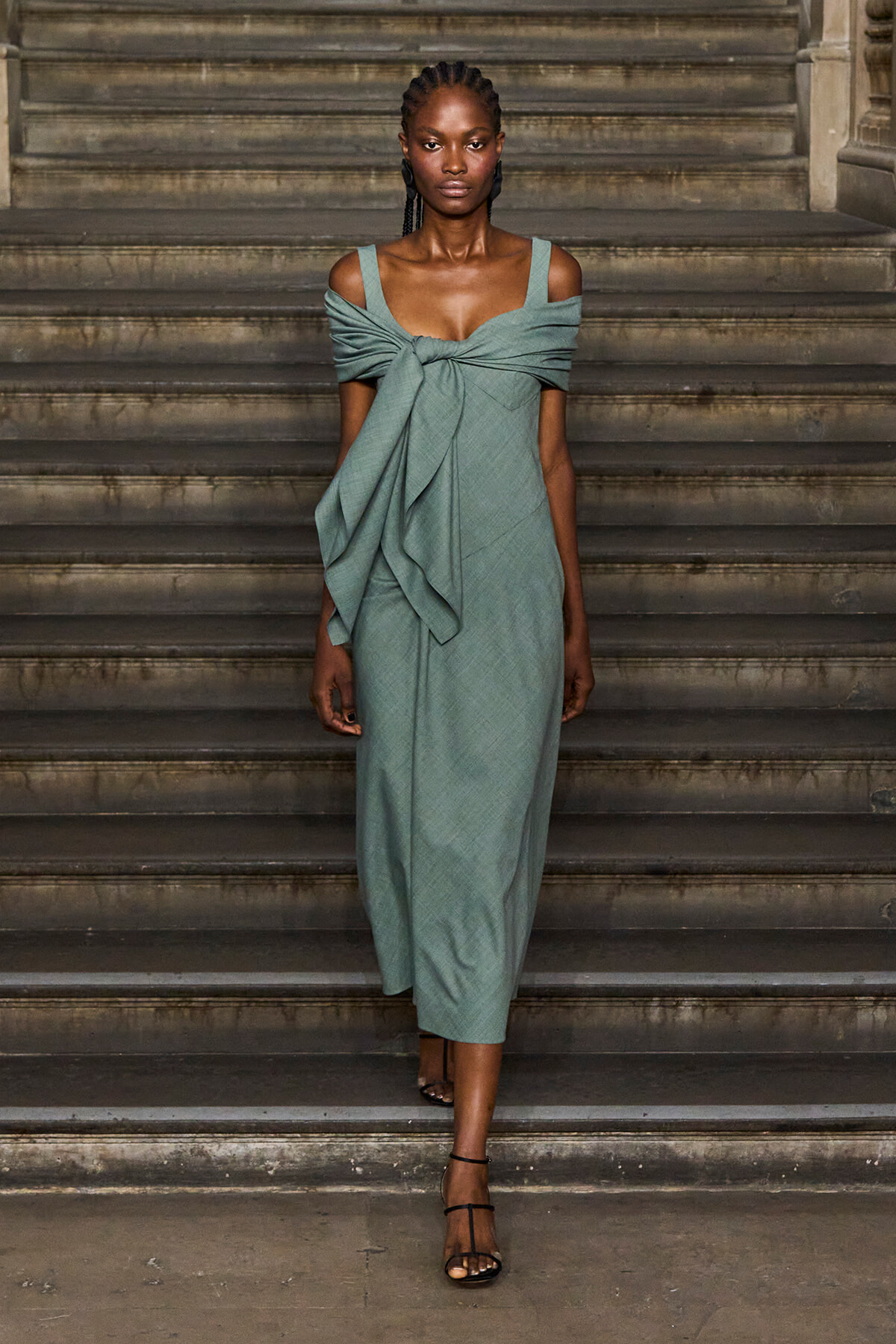 Model in a sage-green off-the-shoulder dress with a front knot standing on stone steps.