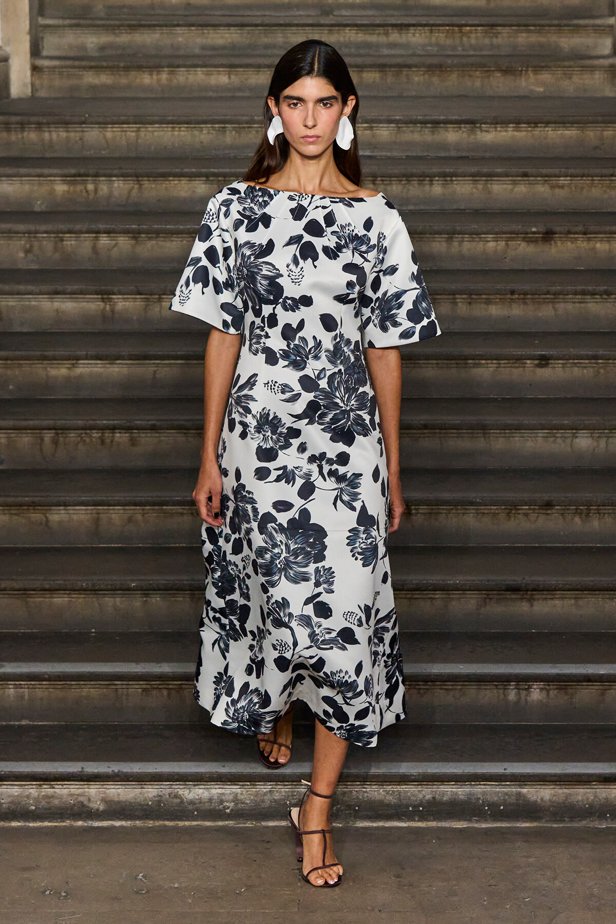 Model wearing a white midi dress with a navy floral print, standing on stone steps with white sculptural earrings.