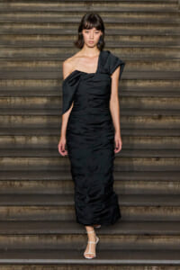 Model wearing a black one-shoulder ruched evening gown, with white strappy sandals, standing on a stone staircase.