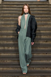 Fashion model on stairs wearing a sage-green wide-leg suit with a black oversized jacket draped over one shoulder, barefoot and minimalist accessories.