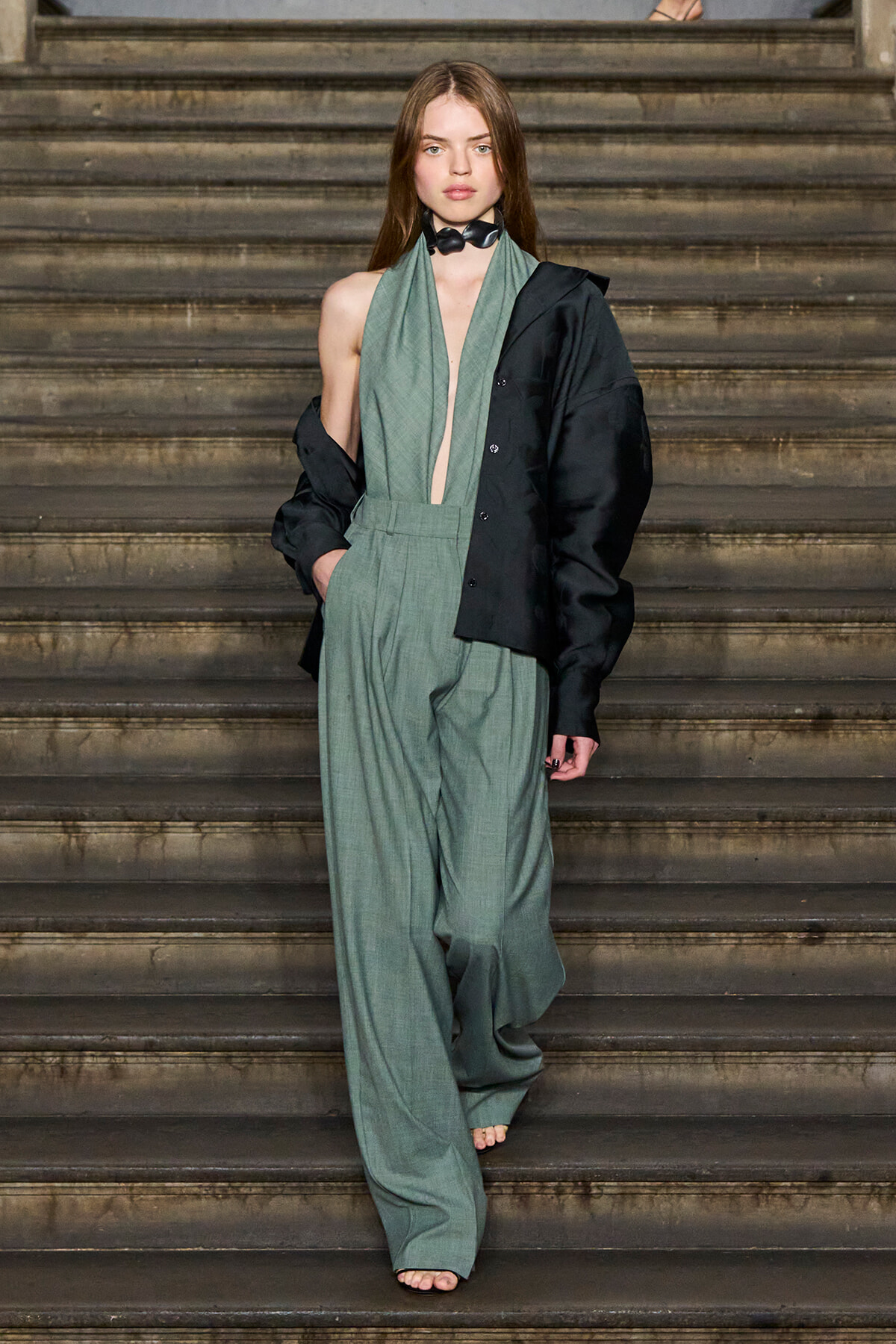 Fashion model on stairs wearing a sage-green wide-leg suit with a black oversized jacket draped over one shoulder, barefoot and minimalist accessories.