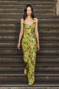 Model walking down stairs in a green-yellow floral jumpsuit with sandals on a weathered stone staircase.
