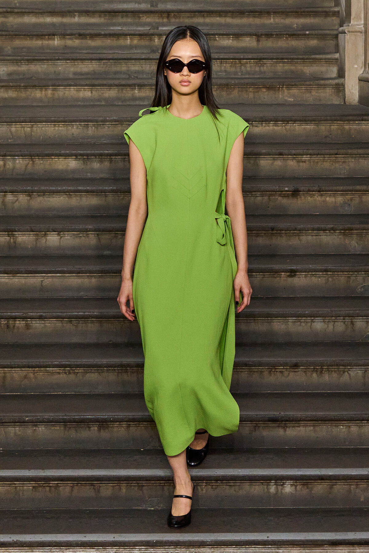 Model in a lime-green dress walking down stone steps, wearing black sunglasses and black heels.