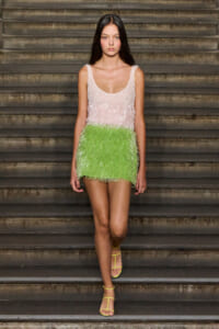 Model wearing a pink-to-green fringe mini dress on stairs with yellow strappy sandals on a runway-like setting.