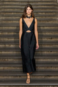 Female model walking down stone stairs in a black sleeveless jumpsuit with a deep plunge neckline and waist cutout, wearing a black petal necklace.