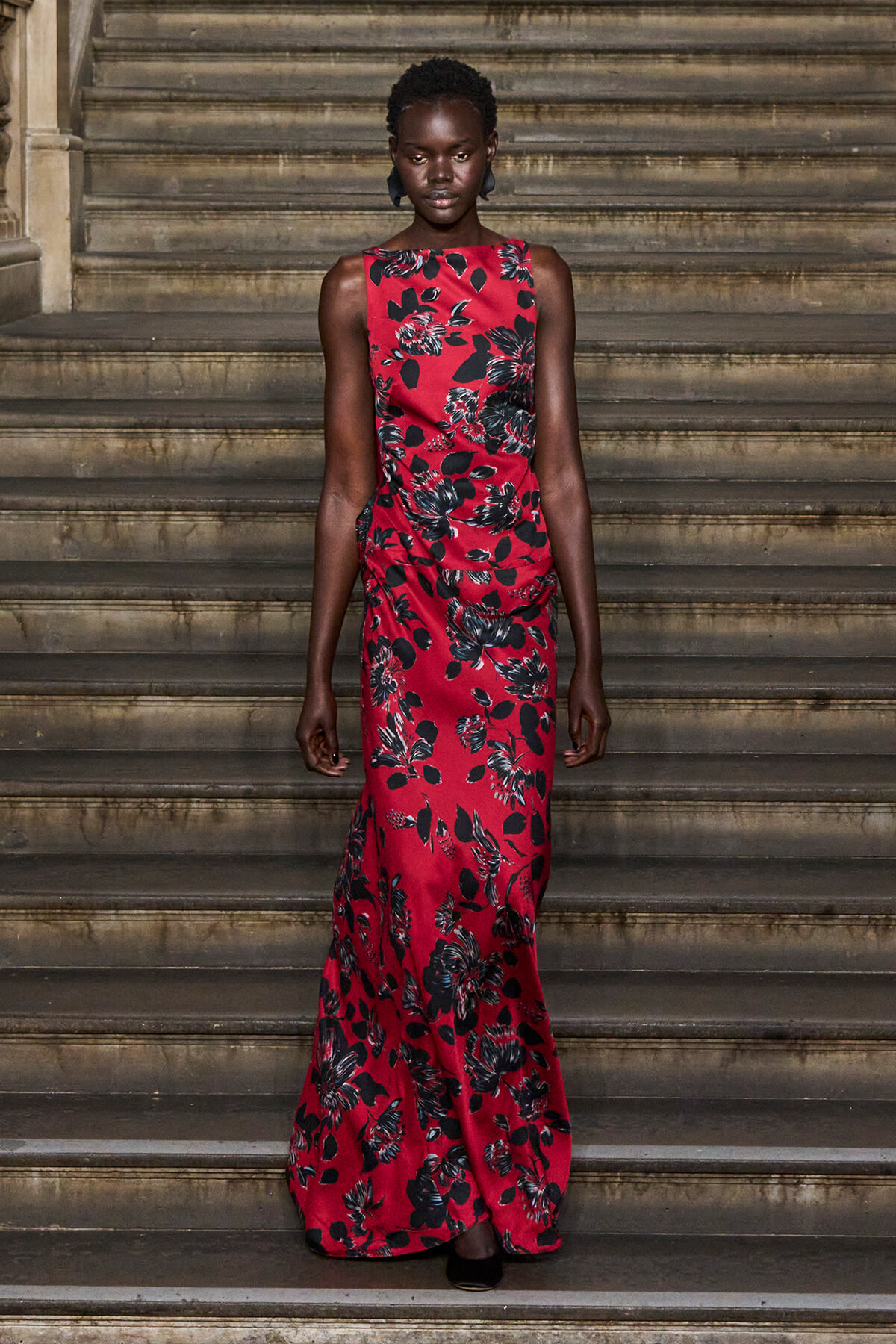 Full-body portrait of a Black woman in a red floral maxi dress standing on stone stairs, facing forward.