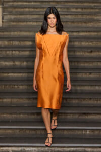 Woman in an orange satin knee-length dress with structured panels walking down concrete stairs in black strappy sandals.