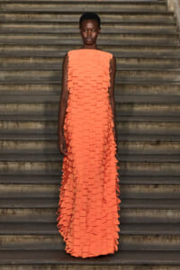 Full-length shot of a woman in a coral-orange, fringe-dressed gown standing on stone stairs.