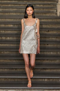 Model walking down stairs in a silver sequined mini dress with orange strappy heels on a runway step backdrop.