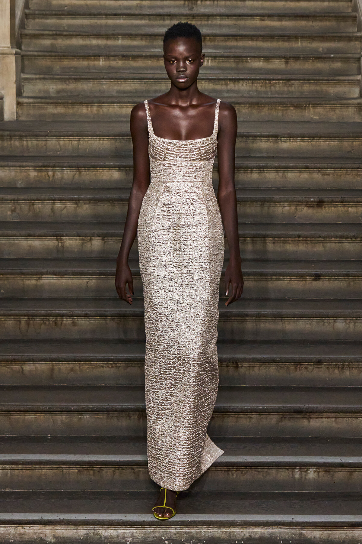 Model wearing a champagne-sequin, floor-length gown on stone stairs with yellow sandals visible at the bottom.