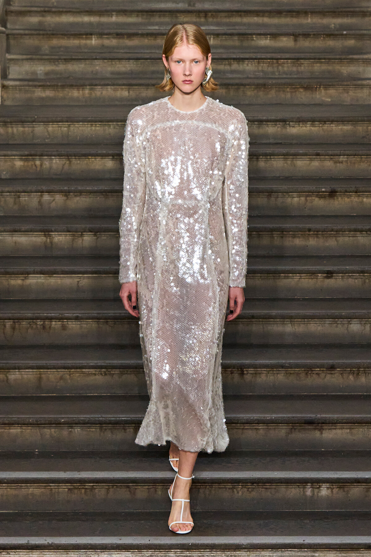 Model wearing a full-length silver sequin dress with long sleeves on a stairway.