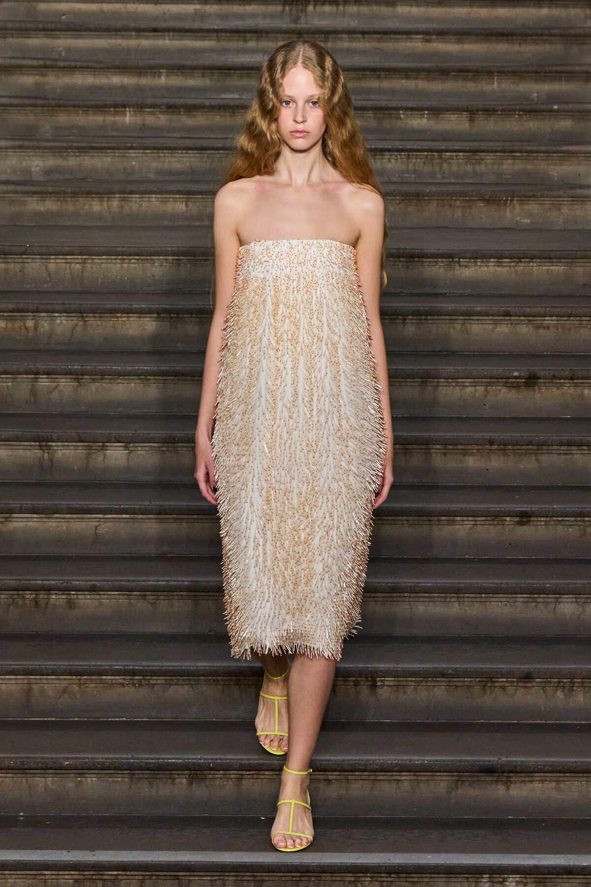 Female model wearing a strapless cream fringe dress standing on dark stairs with yellow strappy sandals.