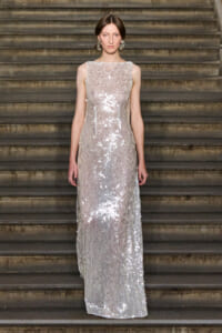 Woman in a floor-length silver sequin gown standing on a stone staircase, facing the camera with a neutral expression.