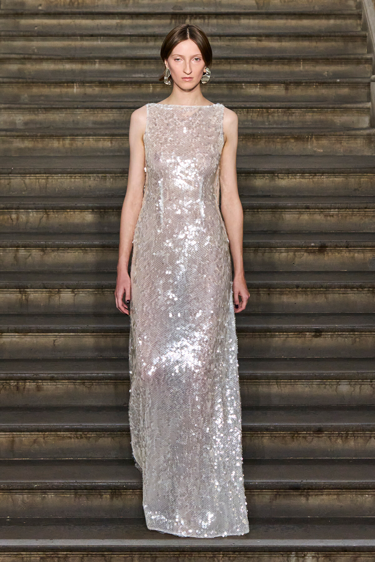 Woman in a floor-length silver sequin gown standing on a stone staircase, facing the camera with a neutral expression.