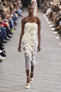 Model walks the runway in a strapless white lace dress with floral appliqués, audience flanking the catwalk.