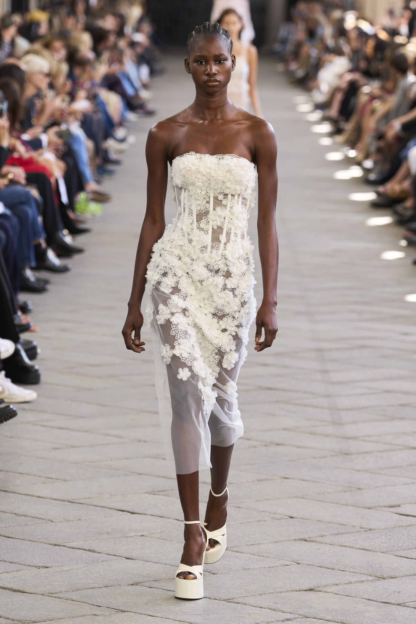 Model walks the runway in a strapless white lace dress with floral appliqués, audience flanking the catwalk.