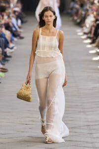 Model walking a runway in a sheer white dress with a ruffled waist, carrying a woven straw bag and wearing strappy sandals as a blurred audience watches behind.