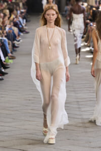 Model walking a fashion runway in a sheer pale outfit with layered necklaces; audience seated along both sides of the runway.