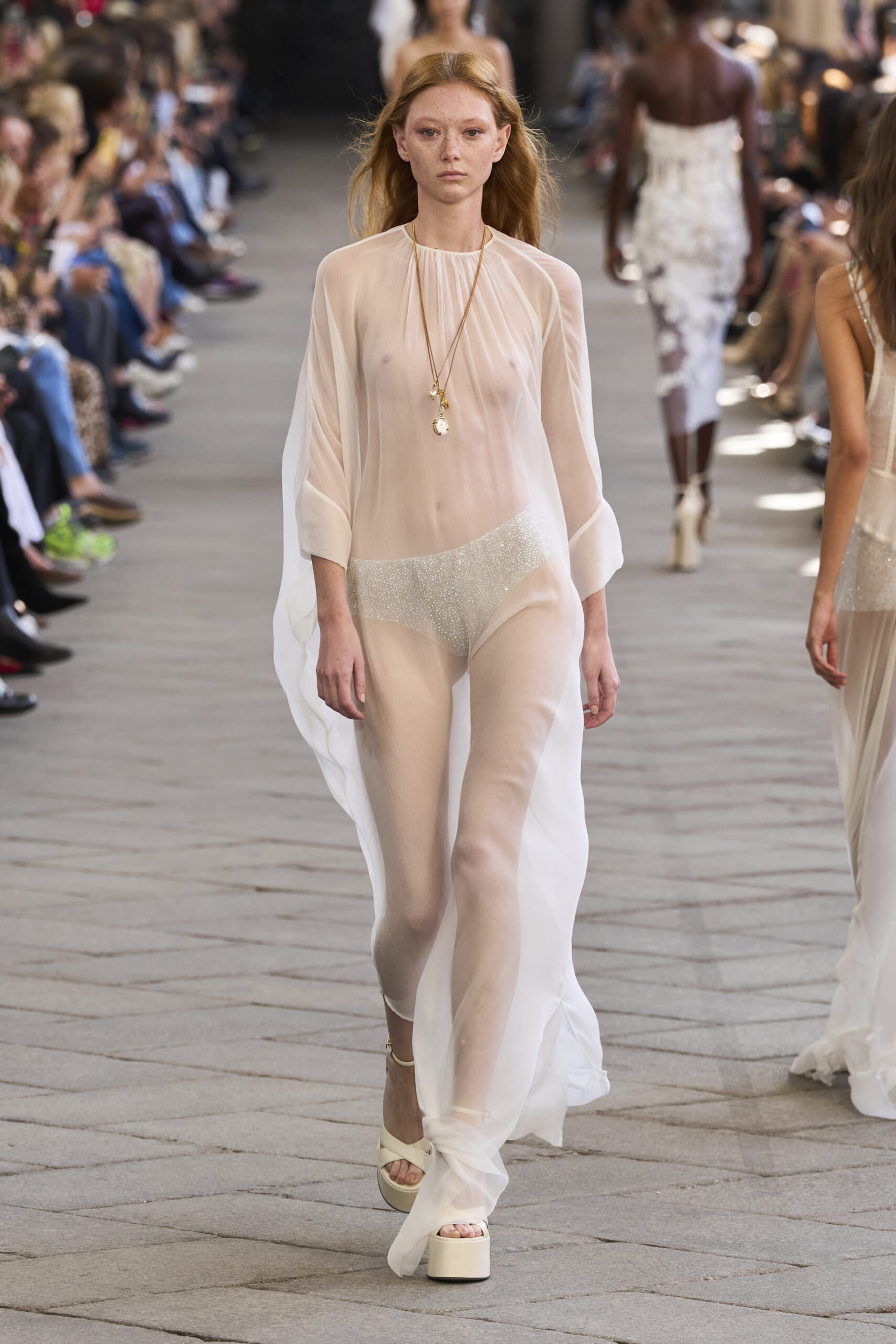 Model walking a fashion runway in a sheer pale outfit with layered necklaces; audience seated along both sides of the runway.
