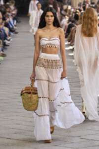 Model walks the runway wearing a white embroidered two-piece boho outfit with a woven straw bag as spectators watch in the background.