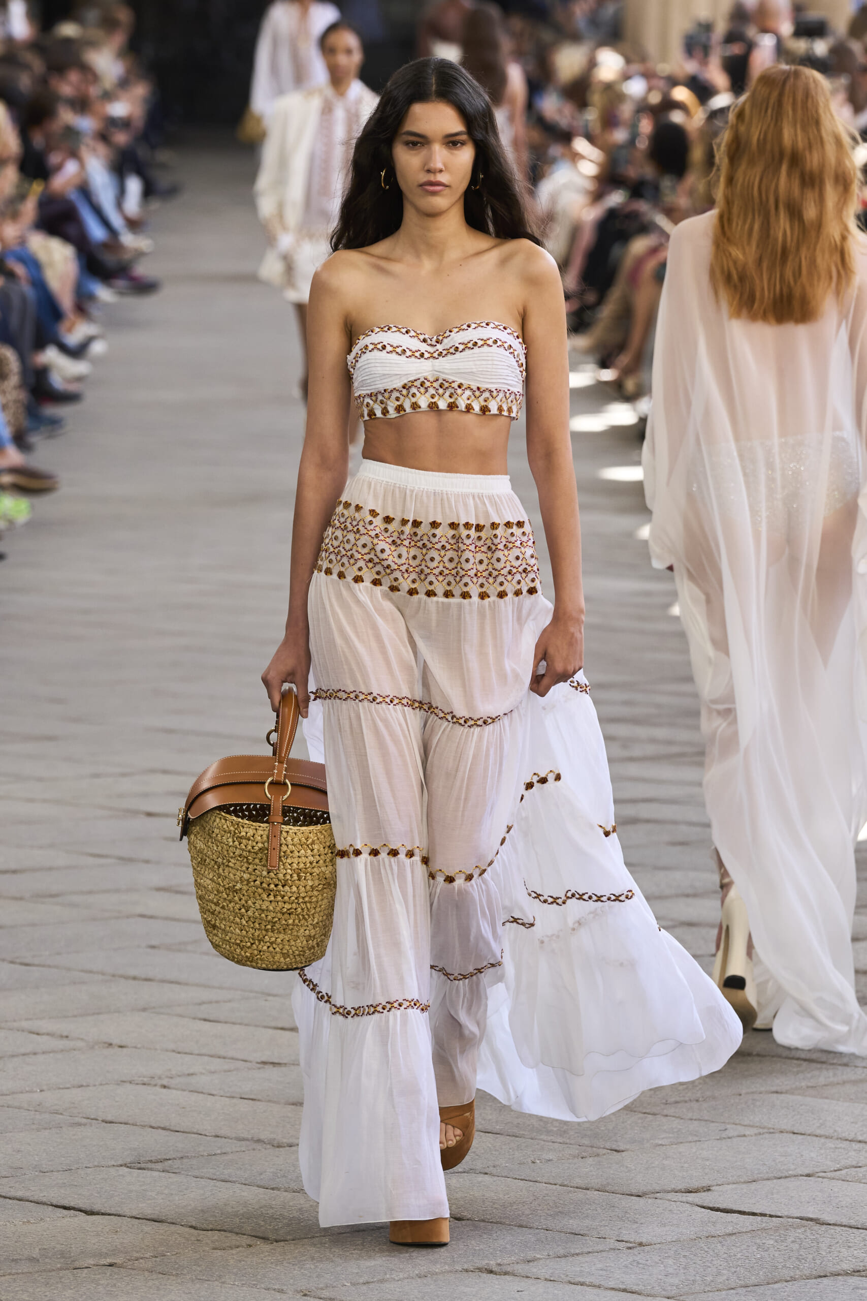 Model walks the runway wearing a white embroidered two-piece boho outfit with a woven straw bag as spectators watch in the background.