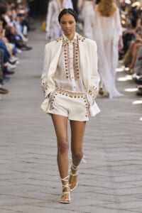 Model walking the runway in a white embroidered suit with brown patterned trim and matching shorts.