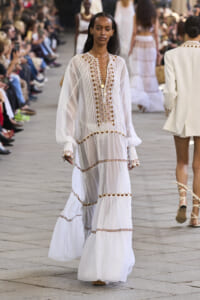 Model walking a fashion runway in a long white embellished dress with gold embroidery along the front and sleeves, audience visible on both sides.
