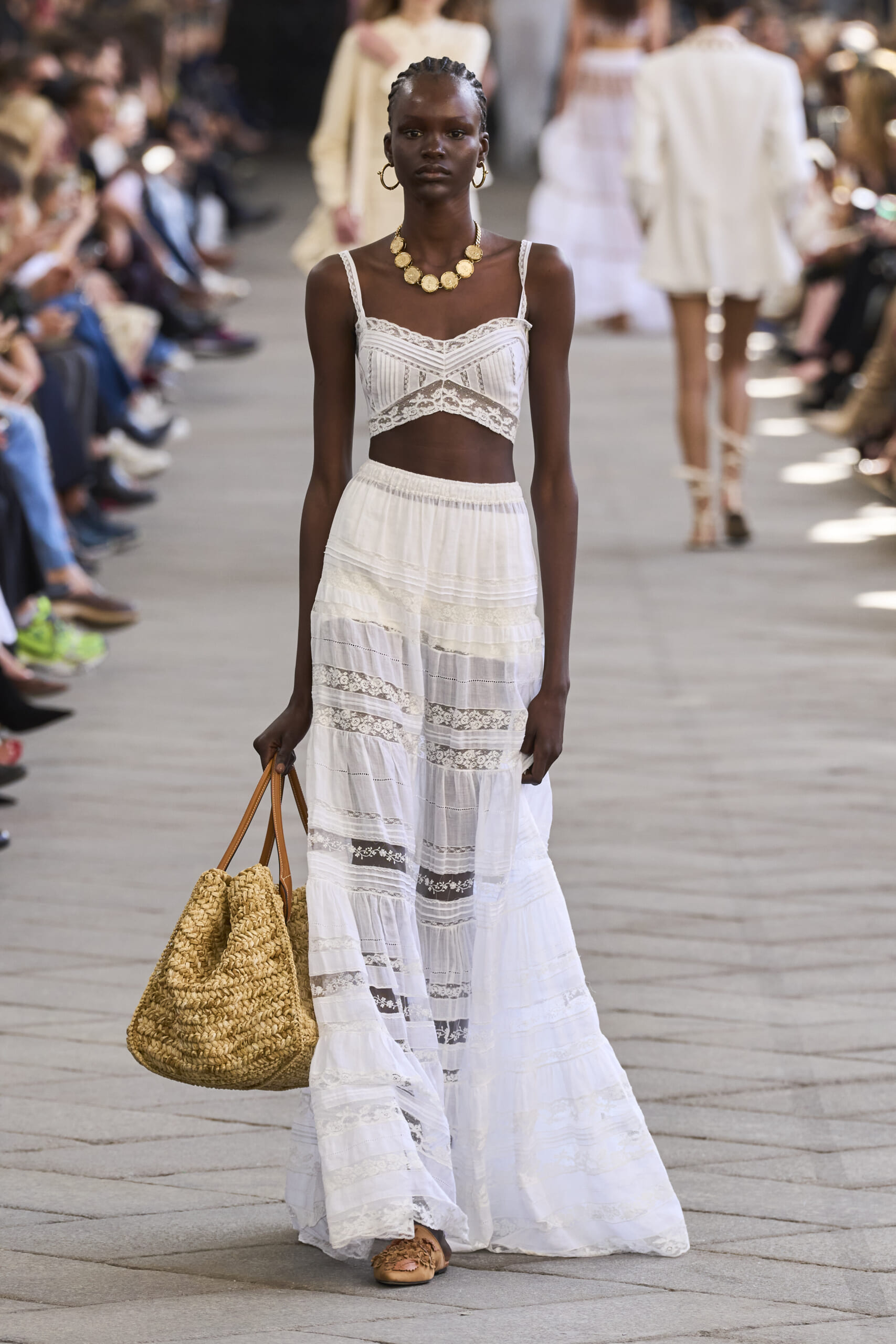 Model walking a runway in a white lace crop top and long tiered skirt, with braided hair, gold hoop earrings, a chunky gold necklace, and a large woven straw bag