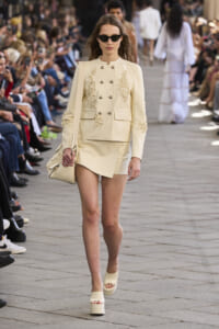 Model walks a runway in a cream embroidered blazer and matching mini skirt, carrying a cream bag and wearing platform sandals.