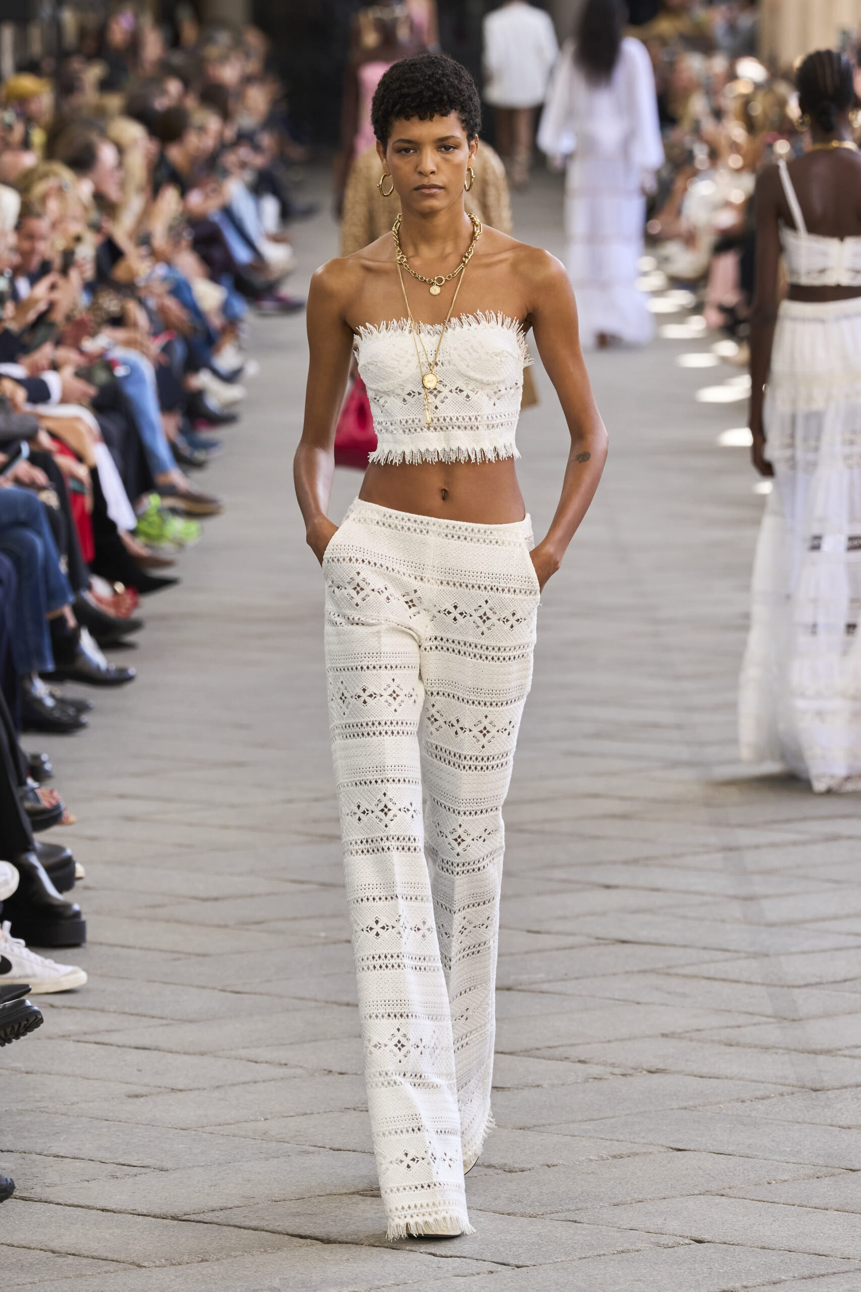 Model walks a fashion show in a white crochet two-piece set with gold jewelry on a crowded runway.