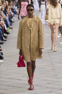 Model walking a runway in a beige embossed coat, pink sandals, and a pink bag, wearing dark sunglasses.