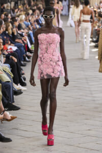 Model walks the runway in a pink floral strapless mini dress with white sunglasses, audience on either side watching the show.