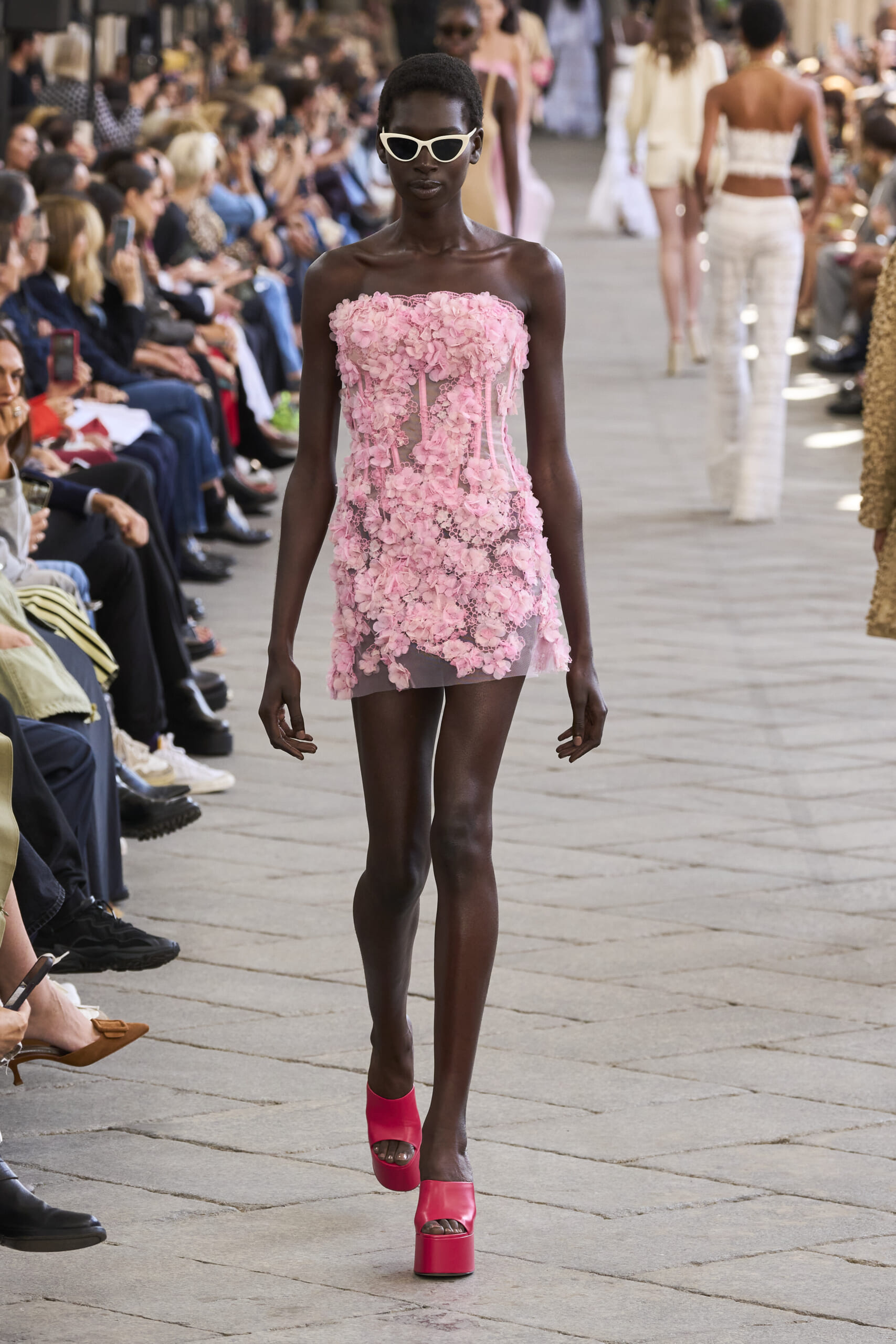 Model walks the runway in a pink floral strapless mini dress with white sunglasses, audience on either side watching the show.