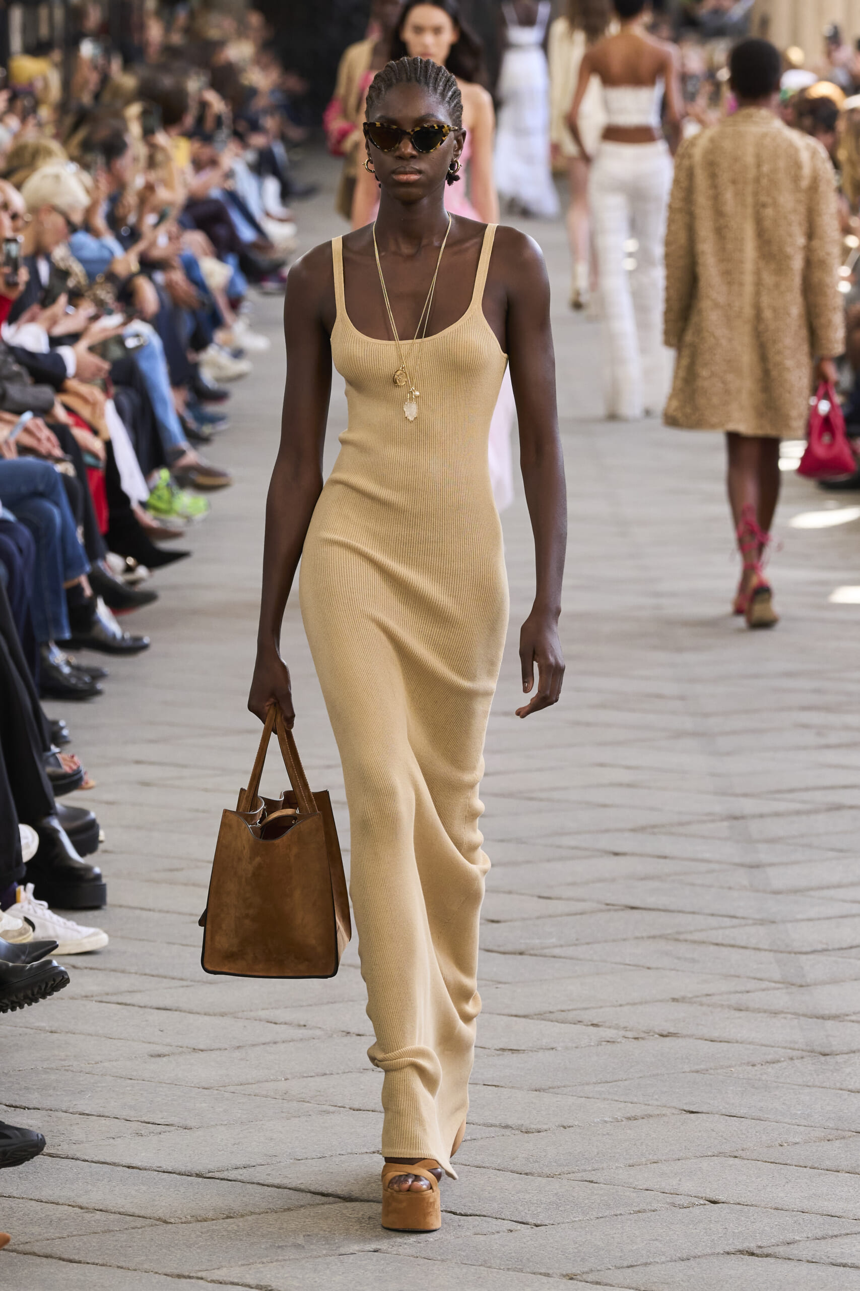 Dark-skinned model walks a fashion runway in a beige ribbed tank dress, sunglasses, layered gold necklaces, and a brown tote bag.