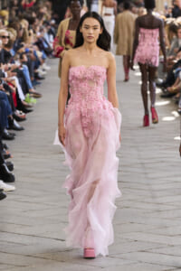 Female model walks a pink, flower-embellished strapless gown down a fashion runway with seated audience on both sides.