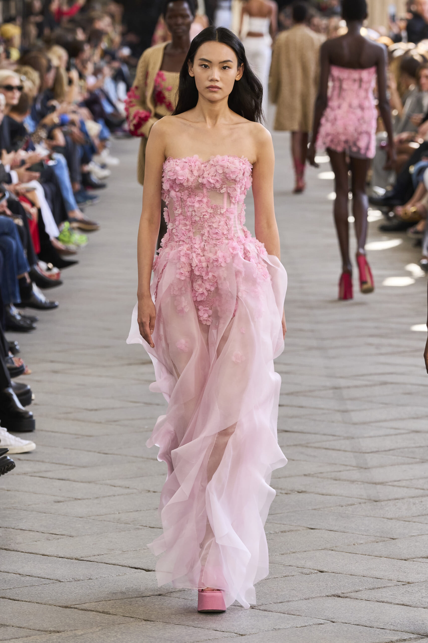 Female model walks a pink, flower-embellished strapless gown down a fashion runway with seated audience on both sides.