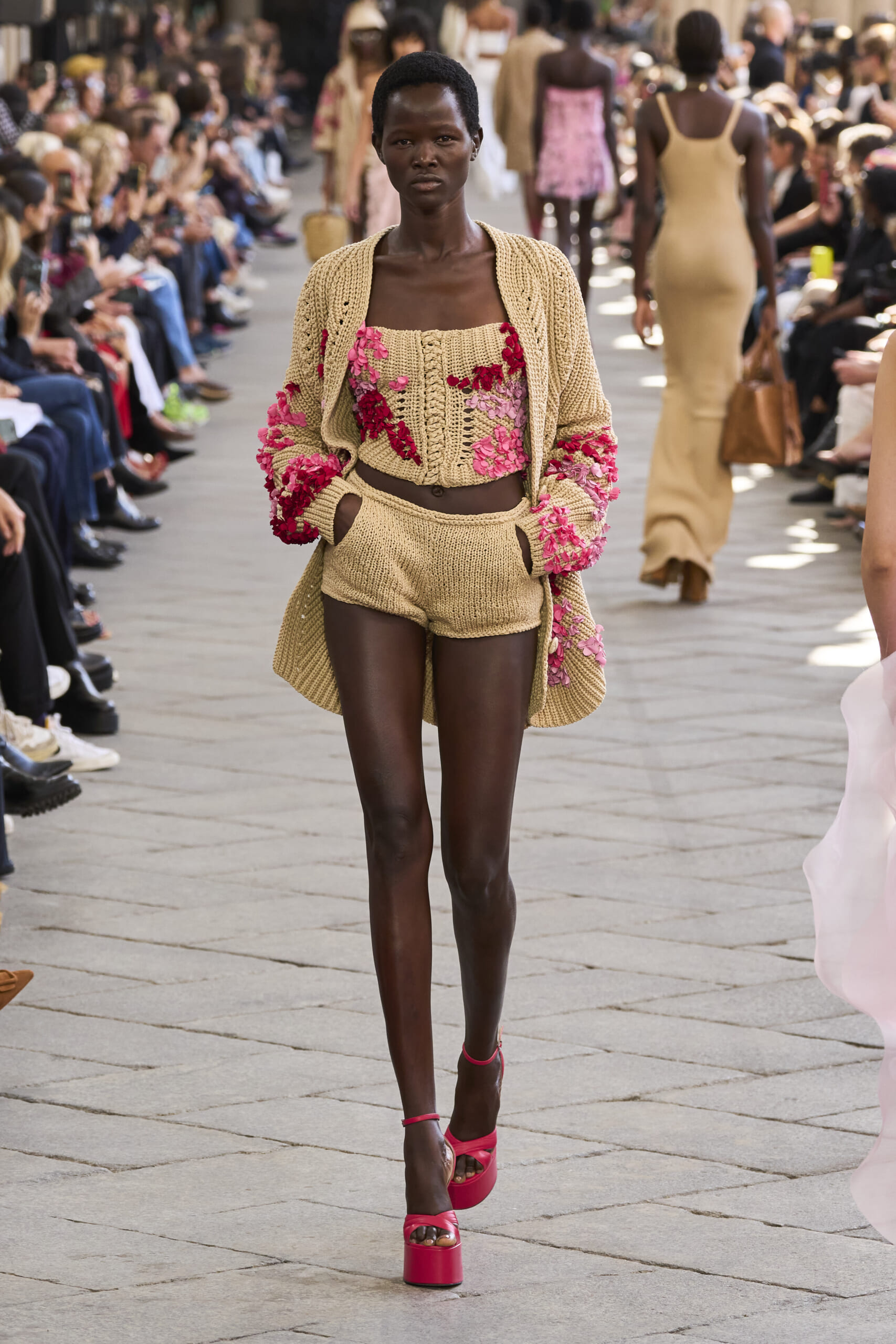 Model walks a fashion show wearing a beige knit set with pink floral embroidery; audience seated along the runway on both sides.