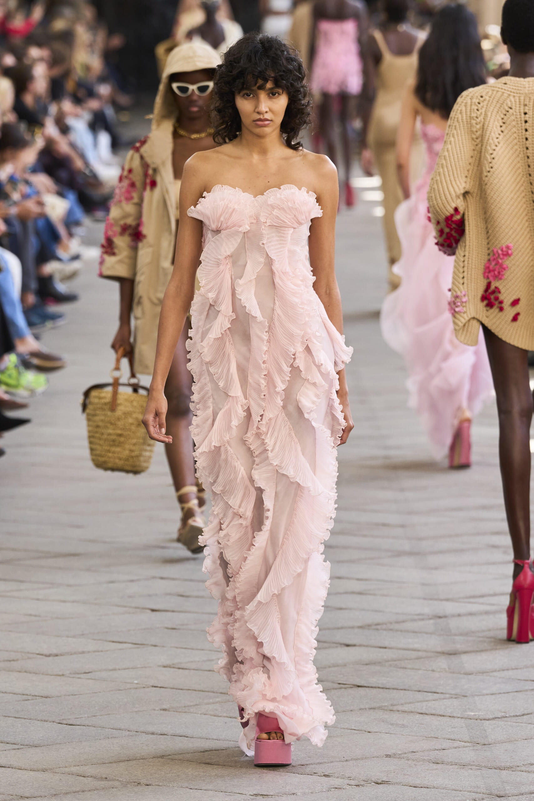 Model walking a fashion runway in a pale pink, ruffled gown with pink platform heels, audience along the sides.