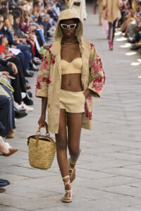 Model on a runway wearing a beige hooded coat with pink floral embroidery, matching bikini top, and shorts, carrying a straw basket bag with sandals visible.