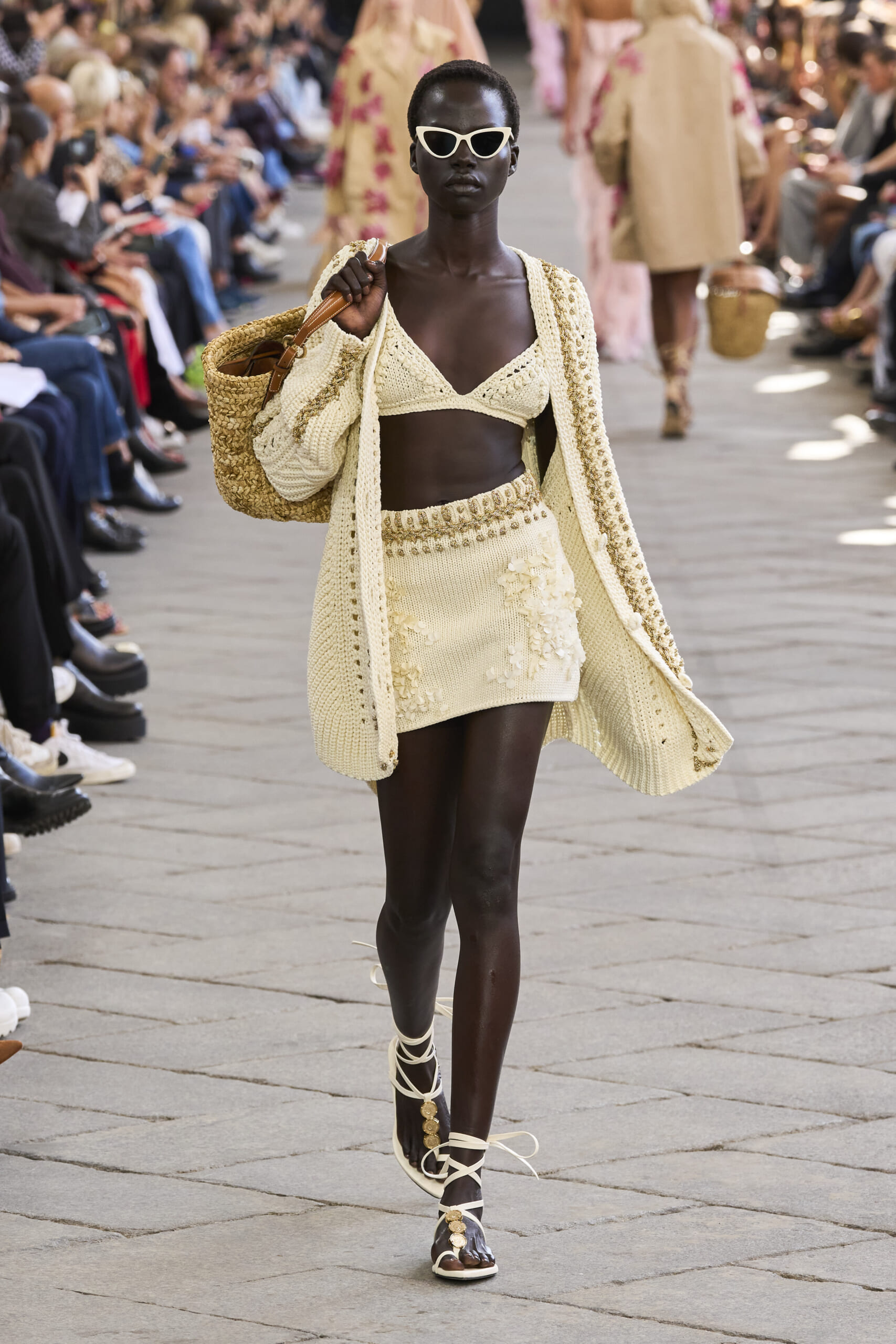 Dark-skinned model walking a runway in a cream crochet outfit with a large woven tote and white cat-eye sunglasses.