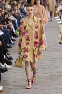 Model walks a runway in a beige coat covered with pink floral embroidery, carrying a matching handbag, wearing red strappy sandals.