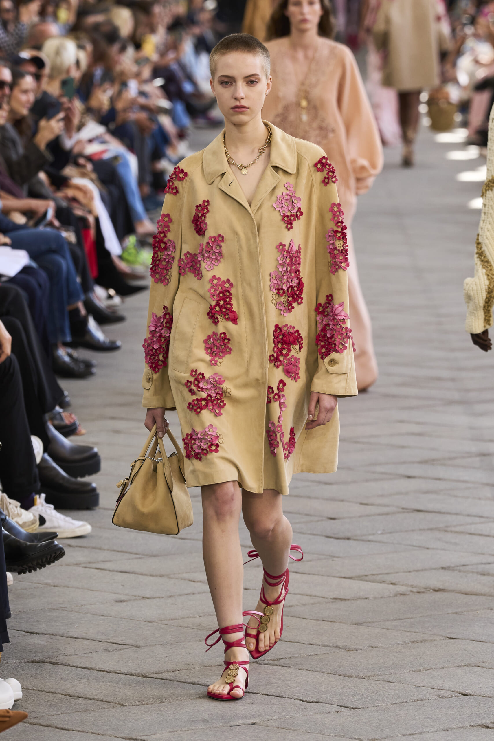 Model walks a runway in a beige coat covered with pink floral embroidery, carrying a matching handbag, wearing red strappy sandals.