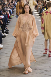 Model walks the runway in a peach satin dress with lace appliqué on the bodice, audience seated on both sides.