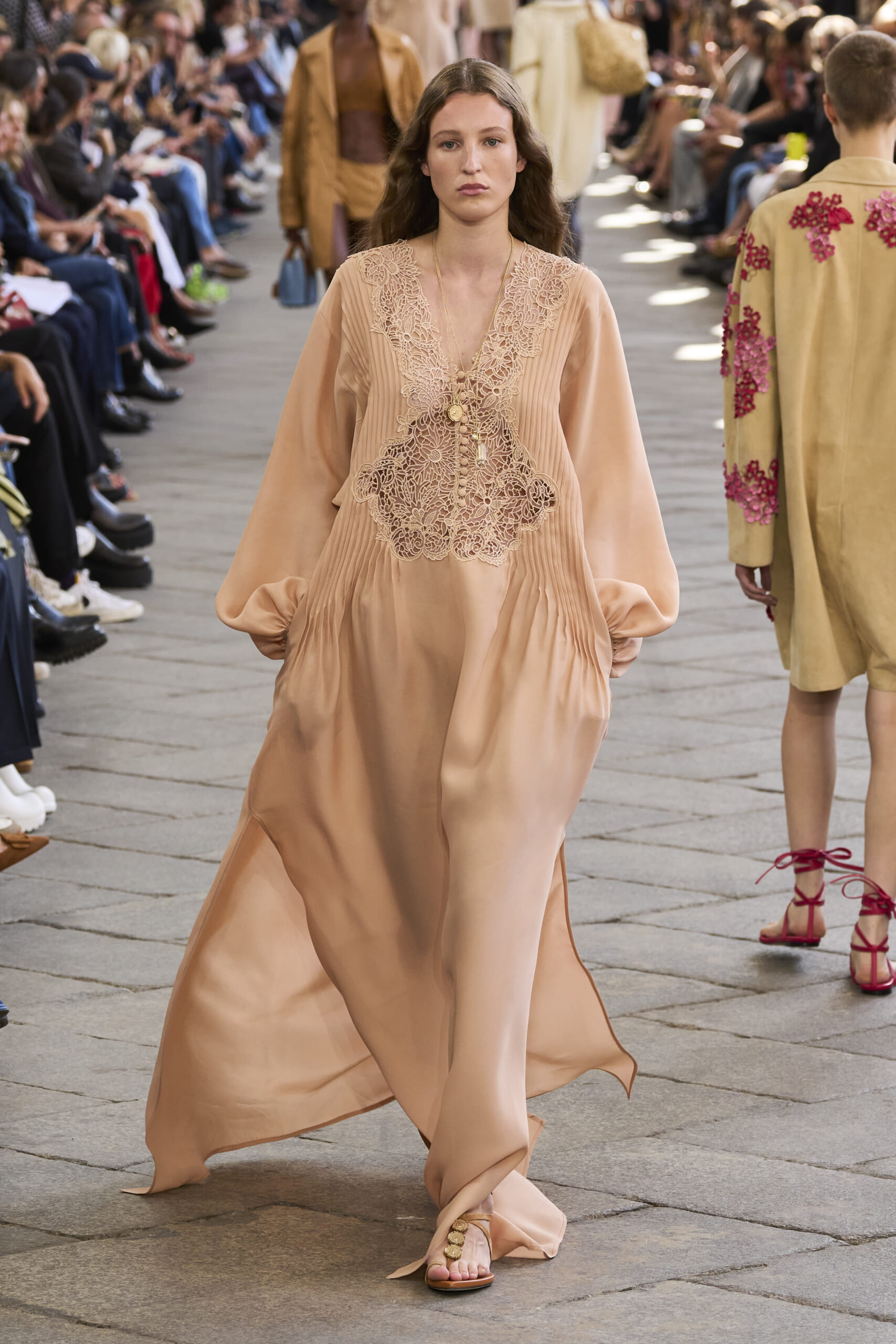 Model walks the runway in a peach satin dress with lace appliqué on the bodice, audience seated on both sides.