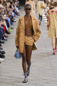Model walks a fashion runway in a tan embroidered coat over matching shorts, wearing dark sunglasses and blue strappy sandals, audience along the catwalk.