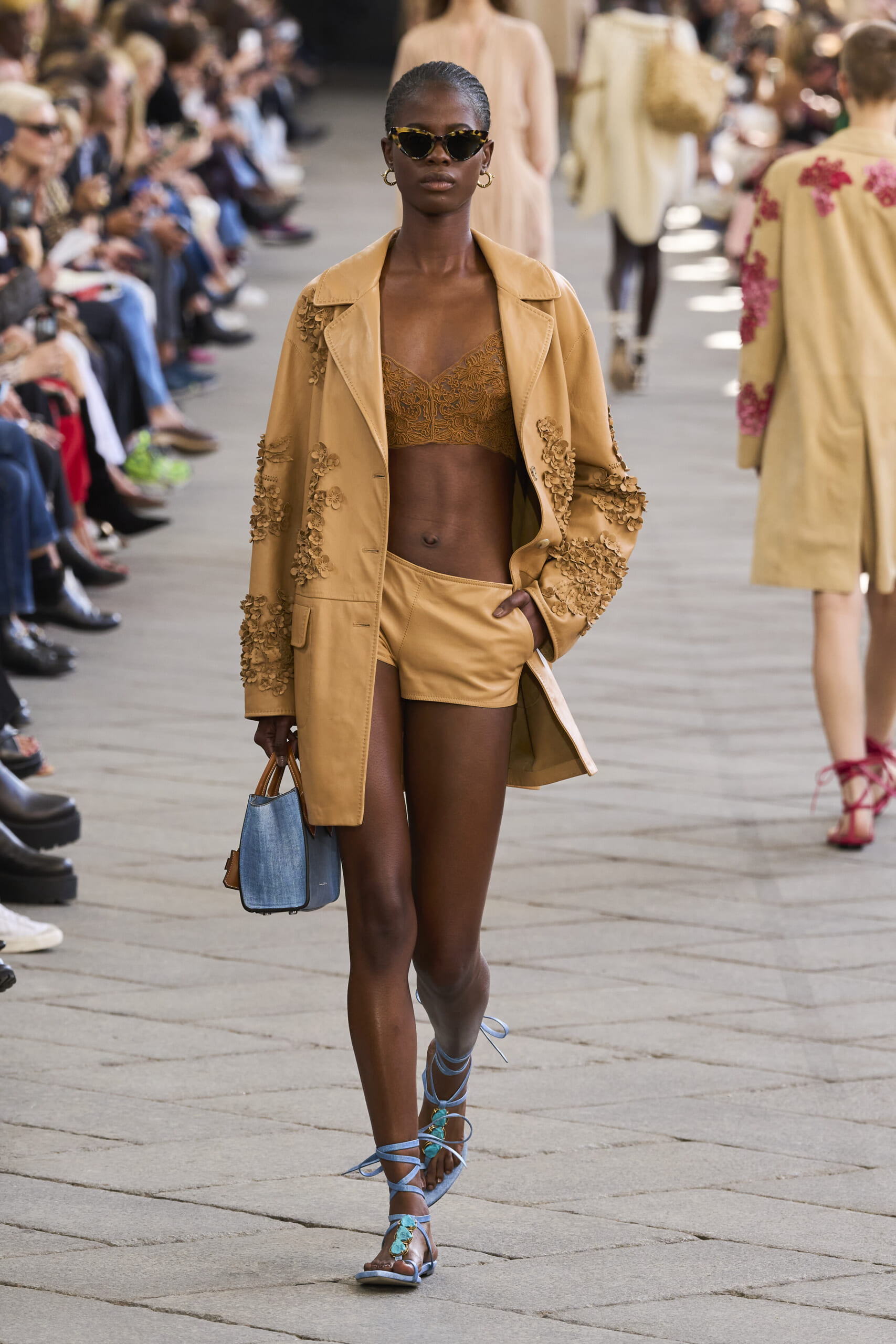 Model walks a fashion runway in a tan embroidered coat over matching shorts, wearing dark sunglasses and blue strappy sandals, audience along the catwalk.