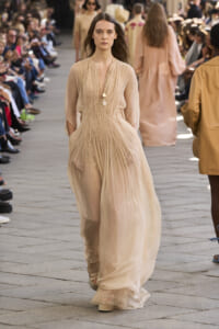 Model walks the runway in a peach pleated gown, audience seated along the sides of the catwalk.