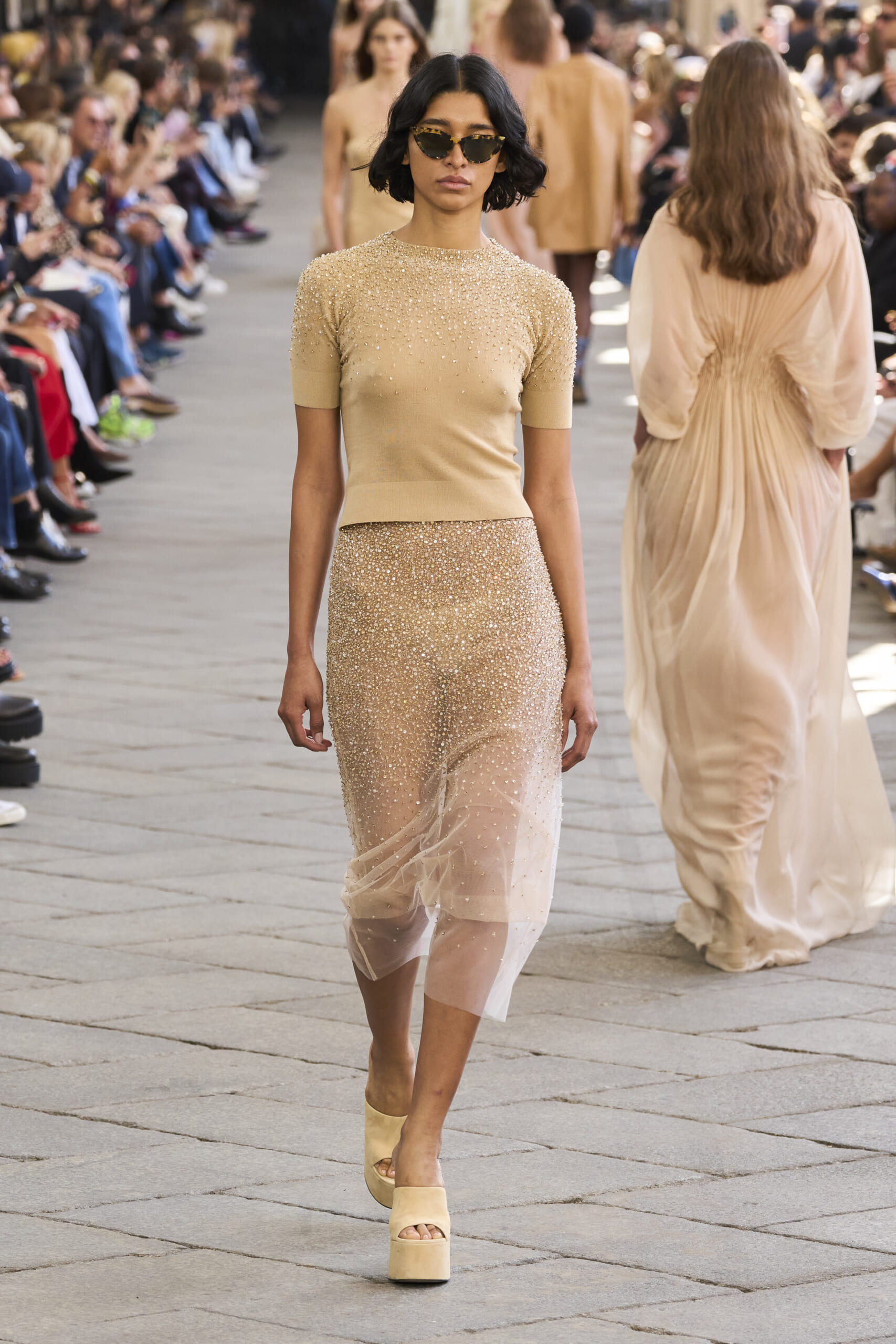 Model walking a fashion runway in a beige beaded dress with sunglasses, audience on both sides.