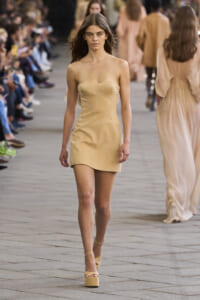 Model walking a fashion show runway in a nude-beige strapless mini dress and beige platform sandals, with a blurred audience in the background.